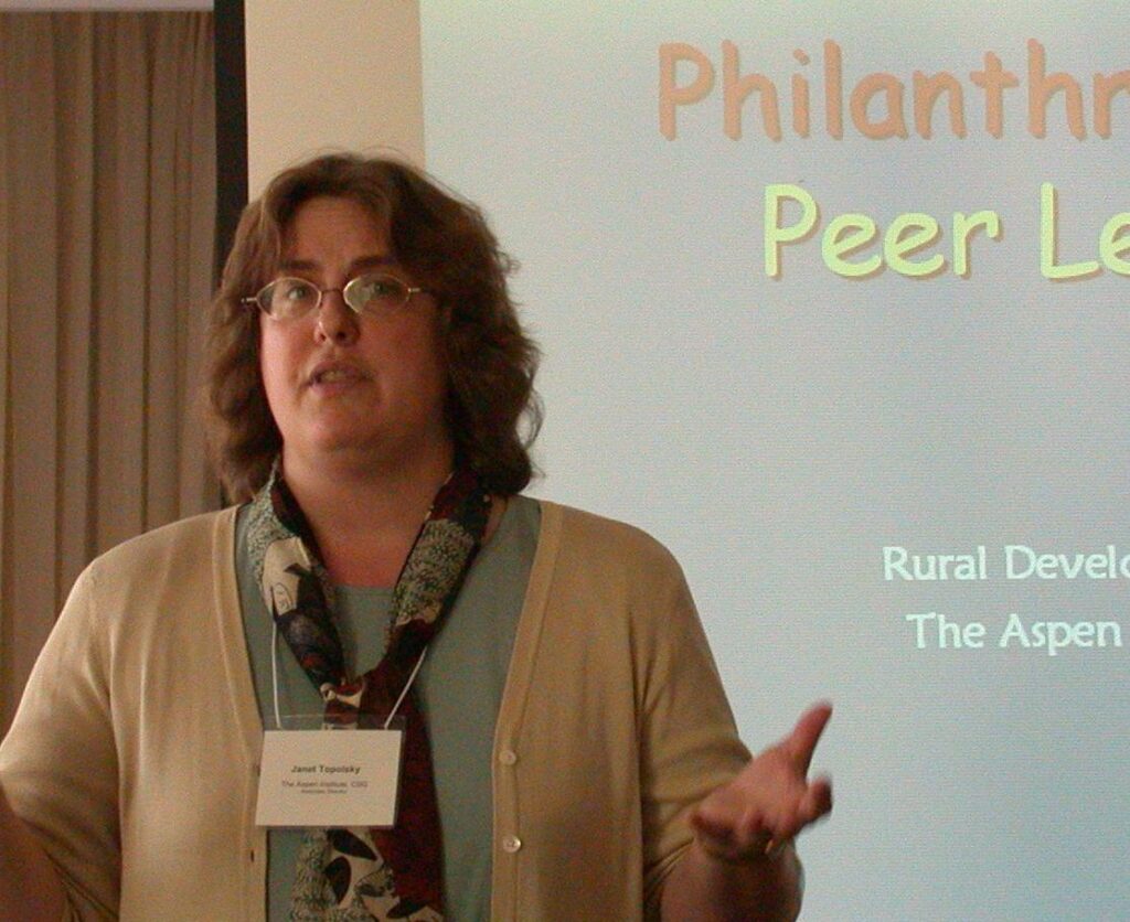  Janet Topolsky speaks at a podium with a presentation slide behind her, leading a Rural Development Peer Learning session as Aspen CSG’s Co-Director.