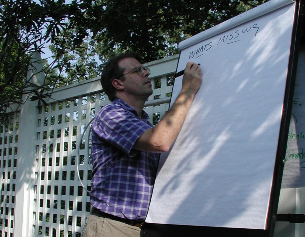 Outdoors in a garden-like setting, a man writes on a large easel pad as part of a brainstorming activity, surrounded by greenery and lattice fencing.
